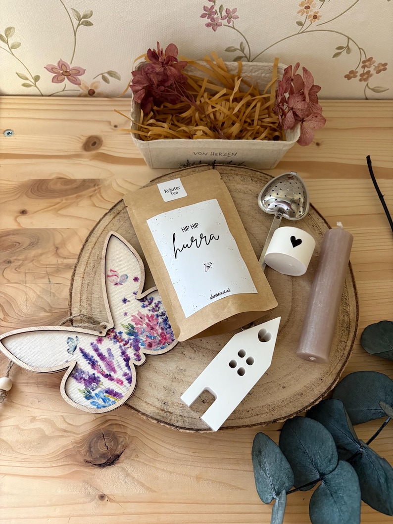 May include: A collection of decorative items on a wooden surface. Includes a butterfly-shaped wooden ornament with a floral design, a bag of tea with the text "HIP HIP hurra", a heart-shaped tea infuser, a candle, and a small white house-shaped object.