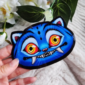 Puede incluir: Una pegatina de gato azul con un diseño de dibujos animados. La pegatina presenta grandes ojos amarillos y rojos, afilados colmillos blancos y una boca sonriente. El gato tiene rayas azules y un contorno negro. La pegatina está sujeta por una mano.