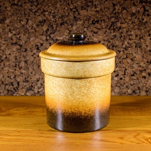 Op de afbeelding: Een bruine en beige keramische pot met een deksel. De pot heeft een gespikkeld glazuur en een donkerbruine band rond de onderkant.