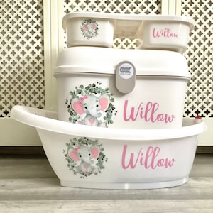 May include: White baby bath set with a pink elephant design and the name "Willow" on each piece. The set includes a large tub, a smaller tub, and a storage container.