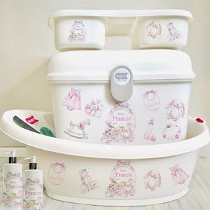 May include: White baby bath set with a large tub, two smaller tubs, and a storage bin. The set features pink and white decals with the name "Baby Francis" and images of baby items like a rocking horse, a bib, and a bottle.