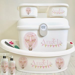 May include: White baby bath set featuring a hot air balloon design. The set includes a tub, a storage bin, two containers, and two bottles. The tub and storage bin have the name "Angela" printed on them. The containers and bottles feature a hot air balloon design with a teddy bear in the basket.