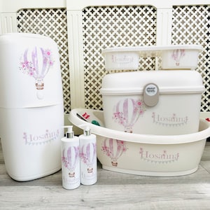 May include: A set of white baby care products featuring a hot air balloon design and the name "Hosanna" printed on them. The set includes a nappy bin, a changing mat, a bath tub, and two bottles of lotion.