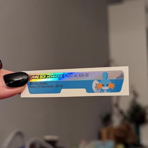 May include: A holographic sticker for a Game Boy Advance with the text "GAME BOY ADVANCE MODEL NO. AGB-101" and "Game Boy Advance Rating: 3.7V 60mA Battery: 2AA LSV". The sticker features a blue cartoon character with orange fins.