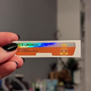 May include: A Game Boy Advance sticker with a holographic top section and an orange bottom section. The sticker features the text "GAME BOY ADVANCE" and a cartoon Torchic character. The sticker also includes technical specifications.