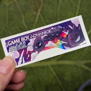 May include: A rectangular sticker with the text "GAME BOY ADVANCE SP" in white and purple. The sticker features cartoon characters on a purple and white background. Model and battery information is also included.