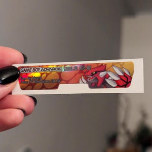 Peut inclure: Un autocollant holographique pour une console Game Boy Advance. L'autocollant présente un fond rouge et orange avec un personnage Pokémon rouge et noir. L'autocollant indique "GAME BOY ADVANCE MODEL NO. AGB-101 Game Boy Advance Rating: 3.7V 60mA Battery: 2AA 1.5V"