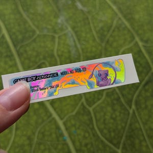 Peut inclure: Un autocollant rectangulaire pour une Game Boy Advance, modèle AGB-101, tenu contre une feuille verte. L'autocollant présente un design coloré avec un personnage de dessin animé violet et le texte "GAME BOY ADVANCE".