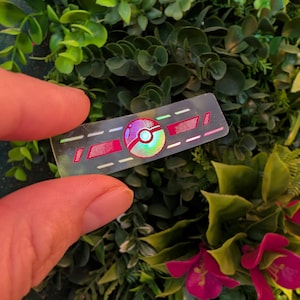 Può includere: Un adesivo rettangolare trasparente con un design olografico. L'adesivo presenta un design centrale rosso e bianco con una sfera colorata e due linee parallele. L'adesivo è tenuto su uno sfondo di fogliame verde e fiori rossi.