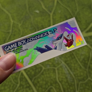 May include: A holographic sticker for a Game Boy Advance SP. The sticker features the text "GAME BOY ADVANCE SP" and a graphic of two characters. The sticker also includes the model number AGS-101 and other technical specifications.