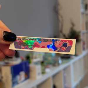 Op de afbeelding: Een holografische sticker voor een Game Boy Advance console. De sticker heeft een roze en blauw ontwerp met een cartoonfiguur, een maan en sterren. De tekst op de sticker luidt "GAME BOY ADVANCE MODEL NO. AGB-101 Game Boy Advance Rating: 32 GumA Battery ZAA1SV".