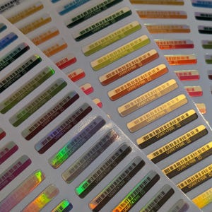 Puede incluir: Hojas de pegatinas rectangulares de colores con códigos de barras y el texto "UG68747666". Las pegatinas tienen un brillo metálico y vienen en varios colores, incluyendo dorado, verde, rojo y azul. Las pegatinas están dispuestas en filas.