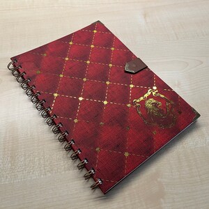 May include: A red spiral bound notebook with a gold lion crest and a gold and red quilted pattern. The notebook has a brown leather closure.