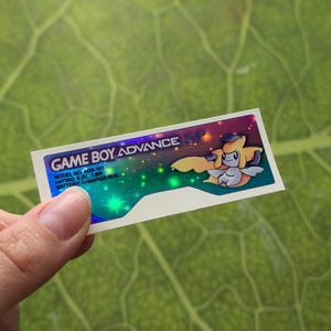 Könnte beinhalten: Ein rechteckiger Aufkleber für einen Game Boy Advance, gehalten von einer Hand. Der Aufkleber hat einen weißen Rand und zeigt den Text "GAME BOY ADVANCE" in Weiß und Lila. Auf der rechten Seite befindet sich eine Cartoon-Figur. Der Hintergrund hat ein farbenfrohes, raumähnliches Design.