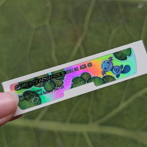 May include: A rectangular sticker for a Game Boy Advance, held by a hand. The sticker has a holographic design with the text "GAME BOY ADVANCE" and "MODEL NO. AGB-101". It features a cartoon character and a green and blue color scheme.