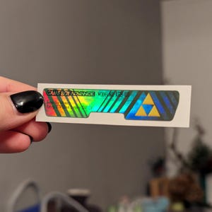 May include: A holographic sticker for a Game Boy Advance with the text "GAME BOY ADVANCE MODEL AGB-101" and a gold triangle logo.