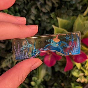 Könnte beinhalten: Ein rechteckiger, transparenter Aufkleber mit einer blauen Cartoon-Figur mit einem orangefarbenen Hut in einer Teichszene. Der Aufkleber hat blaue und goldene Akzente mit blauen Wasser- und Pflanzendetails. Der Aufkleber wird von den Fingern einer Person gehalten.