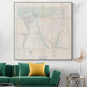 Puede incluir: Un mapa vintage de El Cairo, Egipto, impreso en papel color crema con líneas azules y marrones. El mapa está enmarcado en negro y colgado en una pared blanca sobre un sofá verde con almohadas amarillas.