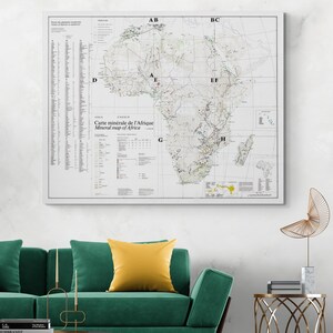Puede incluir: Un mapa mineral de África enmarcado, con texto en francés e inglés. El mapa está detallado con varios símbolos y líneas, sobre un fondo blanco. El marco es blanco y el mapa está colgado en una pared sobre un sofá verde.