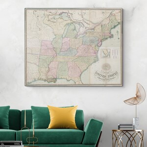 May include: A framed antique map of the United States, displayed on a white wall. The map uses pastel colours and shows detailed geographical information. The words "United States" are visible. The map hangs above a green sofa with a yellow cushion.