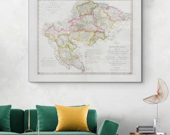 May include: Framed vintage map of Croatia, featuring detailed cartography with various colors highlighting regions and borders. The map is encased in a white frame, displayed above a green sofa with a yellow pillow.