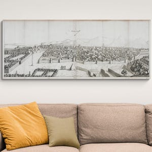 May include: A framed black and white print of a city landscape, likely a historical map or illustration. The artwork depicts a detailed view of buildings, streets, and surrounding mountains. The print is displayed above a sofa with yellow and tan pillows.