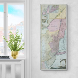 May include: A framed vintage map of the New York and New Jersey area, featuring pastel colors and detailed geographical features. The map is rectangular and vertically oriented, with a light-colored border. It is displayed on a white wall next to a window.