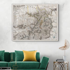 May include: A framed vintage map of the Red Mountain Mining Region in Colorado. The map features detailed topographical lines, text, and a colour palette of beige, grey, and yellow. The map is displayed on a white wall above a green sofa.