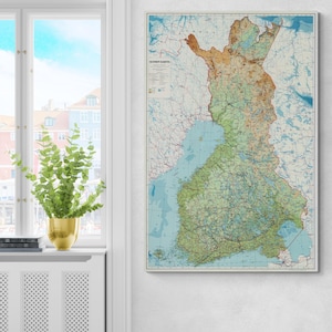 May include: A framed vintage map of Finland, showcasing the country's topography with shades of green, brown, and blue. The map is detailed with roads, lakes, and geographical features, and is displayed on a white wall next to a window.