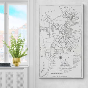 Puede incluir: Un mapa en escala de grises enmarcado de Boston de 1706, con el texto "A Plan of Boston" y el nombre "Ofgood Carleton". El mapa muestra calles, el río Charles y otras características geográficas.