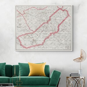 May include: A framed vintage map with a white border. The map features a detailed layout with a prominent red outline highlighting a specific region. The map is displayed above a green sofa with a yellow pillow, creating a contrast of colors.