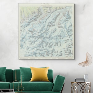 May include: A framed vintage map of Mount McKinley, Alaska, with a detailed topographical view in shades of blue, beige, and white. The map is displayed on a white wall above a green sofa with a yellow pillow. The text "Mount McKinley, Alaska" is visible at the top.