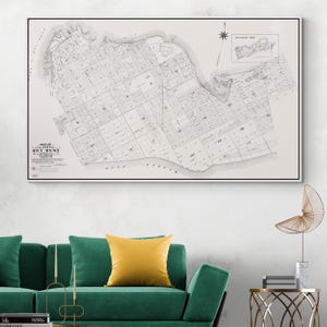 Op de afbeelding: Een ingelijste zwart-wit kaart van Key West, Florida, met gedetailleerde straatindelingen en geografische kenmerken. De kaart bevat de tekst "Map of the City of Key West, Florida". Het kunstwerk wordt weergegeven boven een groene bank met gele kussens.
