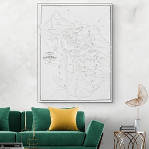 Op de afbeelding: Een ingelijste zwart-wit kaart van de Black Hills regio. De kaart, getiteld "Hutchinson Map Black Hills 1877", toont gedetailleerde geografische lijnen en tekst. Het kunstwerk hangt boven een groene bank met een geel kussen.