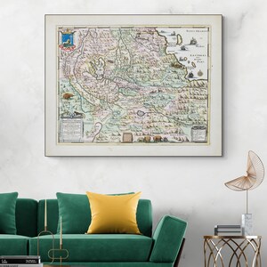 Puede incluir: Un mapa antiguo enmarcado con detalles intrincados y colores vibrantes. El mapa presenta ilustraciones detalladas de formas terrestres, ciudades y texto. El marco es blanco y el mapa se muestra sobre un sofá verde con un cojín amarillo.