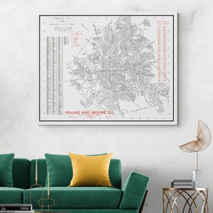 May include: A framed vintage map of Charlotte, NC, with the text "Charlotte, N.C." and "Pound and Moore Co." The map is in black and white with red accents, and is displayed above a green sofa with a yellow pillow.