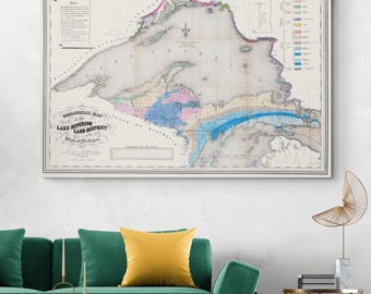 Op de afbeelding: Een ingelijste vintage geologische kaart van het Lake Superior Land District, met gedetailleerde, kleurgecodeerde geologische formaties. De kaart bevat tekst en een schaal in mijlen, met een witte rand en een zwart frame. De kaart wordt weergegeven boven een groene bank met een geel kussen.