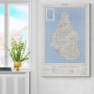 May include: A framed map of Montserrat, a Caribbean island, showing detailed topography. The map includes a grid, roads, and the text "Montserrat". The map is displayed on a white wall near a window, with a plant in a gold pot.
