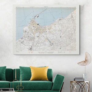 Puede incluir: Un mapa enmarcado de Trípoli, Libia, con el diseño de la ciudad en blanco y negro, sobre un mar azul claro. La palabra "TRIPOLI" está en la parte superior. El mapa se muestra sobre un sofá verde con un cojín amarillo.