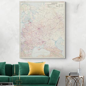 Puede incluir: Un mapa vintage enmarcado de Europa, con una red de carreteras y ciudades en tonos rosas y morados sobre un fondo crema. El mapa se exhibe en una pared blanca sobre un sofá verde con un cojín amarillo.