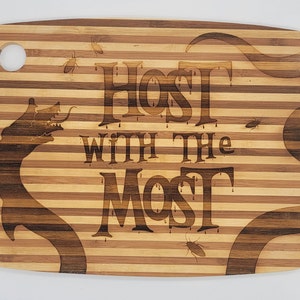 Puede incluir: Una tabla de cortar de bambú marrón y beige con un diseño en blanco y negro de una criatura y el texto "Host with the Most".