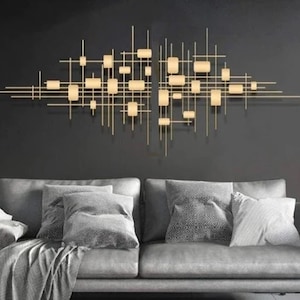 May include: A gold metal wall sculpture with a geometric design. The sculpture is made up of many thin lines and squares, creating a modern and abstract look. It is hanging on a dark gray wall above a gray sofa with pillows.
