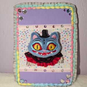 May include: A plush cat face with a top hat and a red and black ruffled collar is centered on a rectangular, multi-layered decorative piece. The background features a polka dot pattern, with a purple border and embellished with rhinestones.