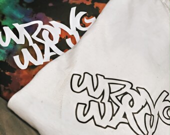 May include: Two t-shirts are shown, one white and one tie-dye. The white t-shirt features the words "WRONG WAY" in black graffiti-style lettering. The tie-dye shirt has the same text in white. The tie-dye shirt has a mix of colours.
