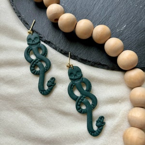 May include: A pair of dark green snake earrings with a skull detail. The earrings are made of polymer clay and have a gold hook.