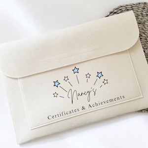 May include: A cream-coloured felt envelope with a flap closure. The front features a rectangular patch with the text "Nancy's Certificates & Achievements" and a starburst design with colourful stars. The envelope is on a white surface.