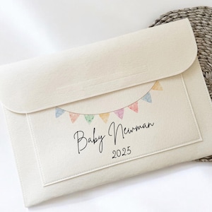 May include: A cream-coloured envelope-style pouch with a flap closure. The front features a watercolour banner of pastel flags and the text "Baby Newman 2025" in a handwritten font. The pouch is on a woven circular mat.