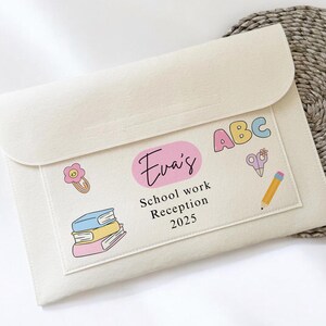 May include: A cream-coloured school work folder with a flap closure. The folder has a pocket with the name "Eva's", the text "School work Reception 2025", and colourful illustrations of books, a flower, scissors, a pencil, and the letters "ABC".