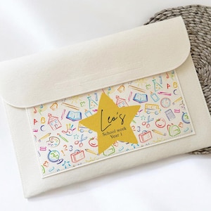 May include: Off-white felt schoolwork folder with a clear pocket displaying a colourful school-themed pattern. A gold star with the name "Leo's" and "School work Year 1" is attached. The folder is on a woven mat.