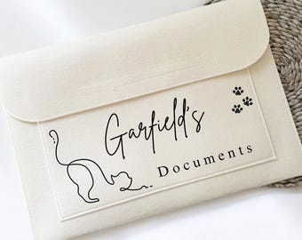 May include: Cream document holder with a flap closure. It features the text "Garfield's Documents", a cat outline, and paw prints. The holder is on a woven circular mat.